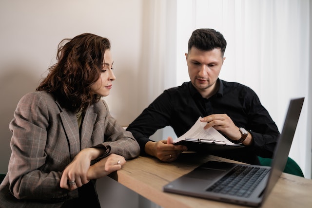 a landlord with curly brown hair in a brown plaid suit reviews a rental agreement with their new tenant who holds a clipboard with a document on it